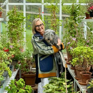 Cath Kidston MBE holding dog in greenhouse filled with pelargoniums and other plants