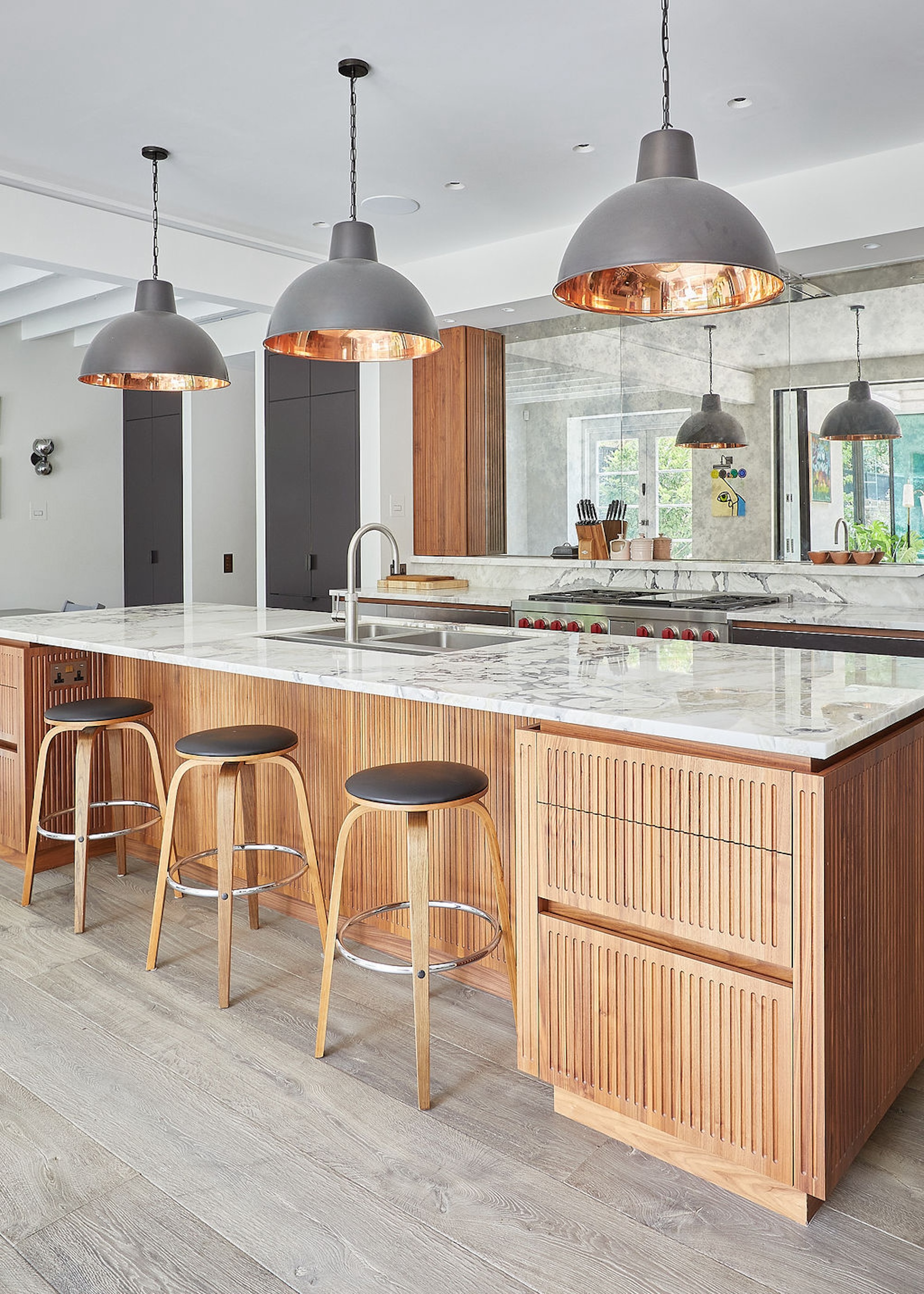 A wood kitchen showing an island with fluted cabinet fronts and a marble worktop with a trio of oversized domed pendant