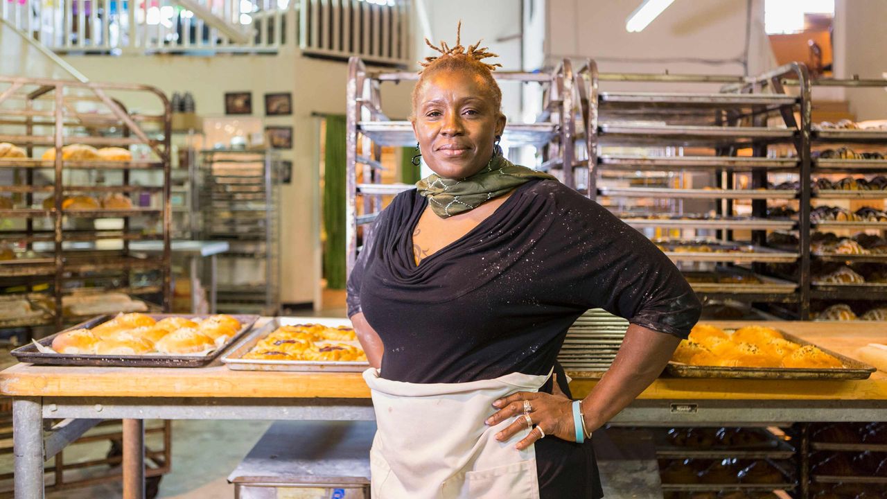 A woman with an apron stands in her bakery with her hands on her hips.