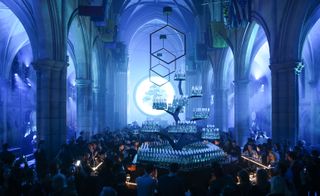 View of a large crowd of people inside The American Cathedral in Paris. The cathedral is lit up an icey blue colour, there is a tall tree display with glass bottles on the branches, hexagon shapes are hanging from the ceiling and a round light display featuring a tree can be seen at the back of the cathedral