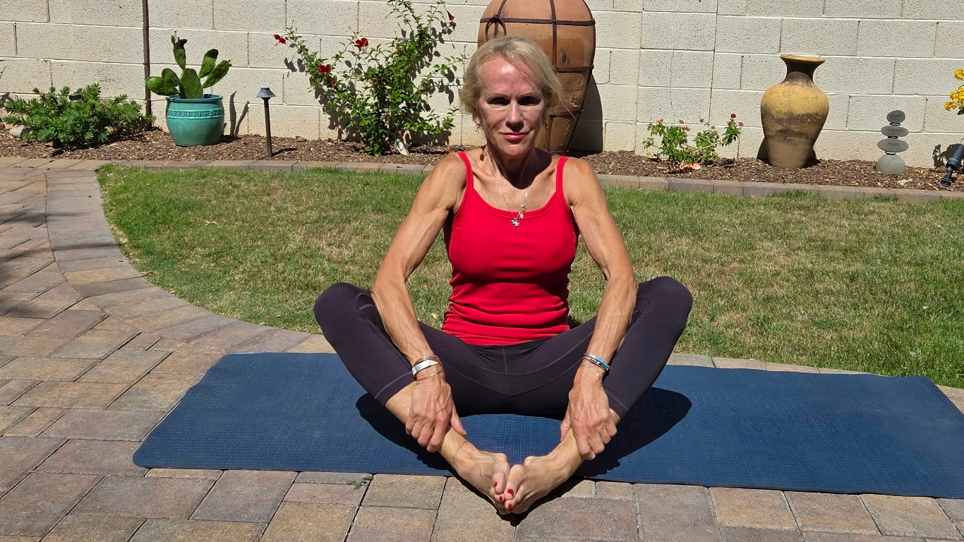 Woman in black leggings and red tank top on exercise mat on patio outside, next to grass.