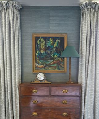 An antique dresser with a vintage clock, a tall lamp and a framed picture hanging above