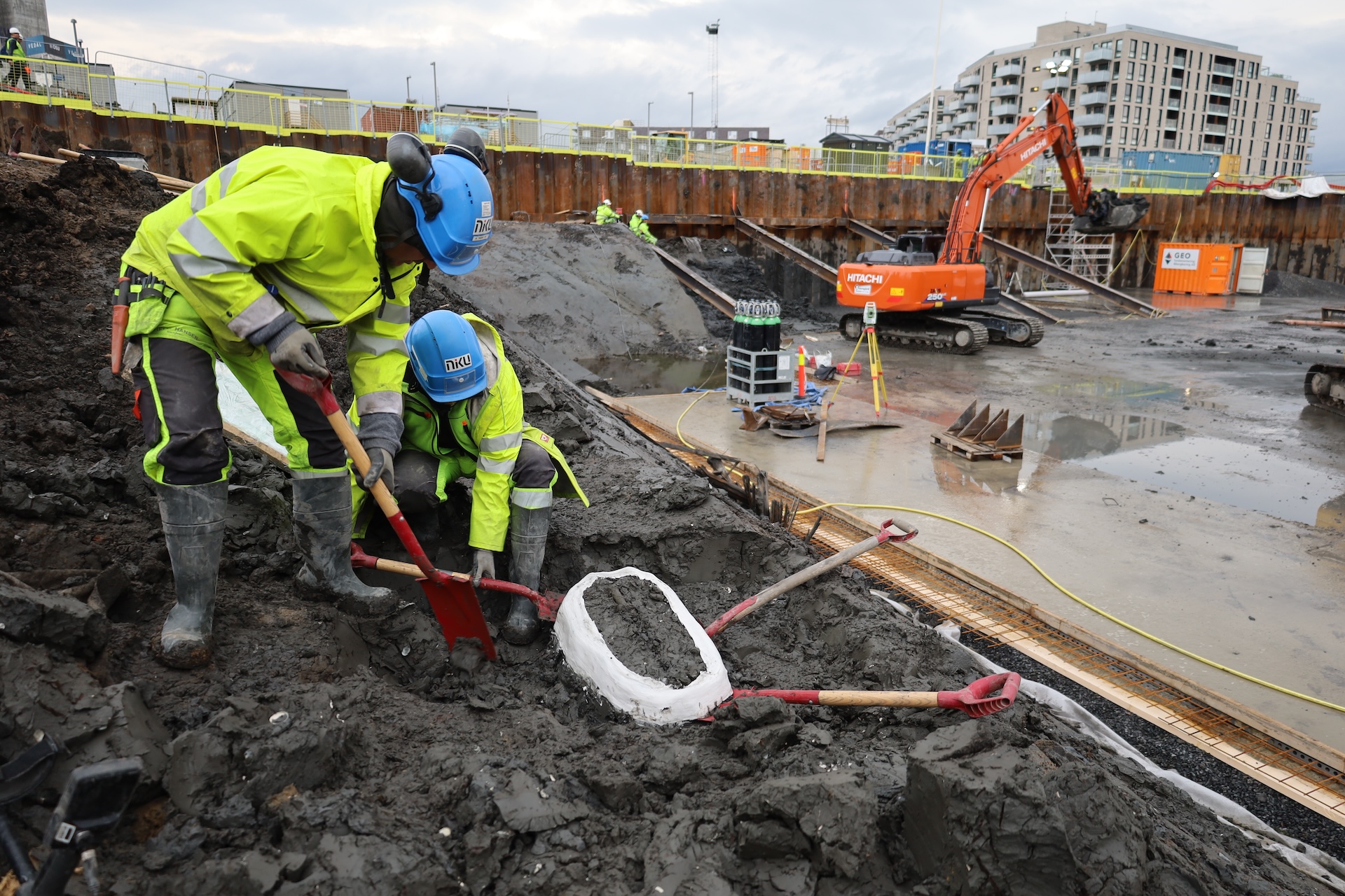 Men excavate around black rocks with Oslo buildings visible in the background