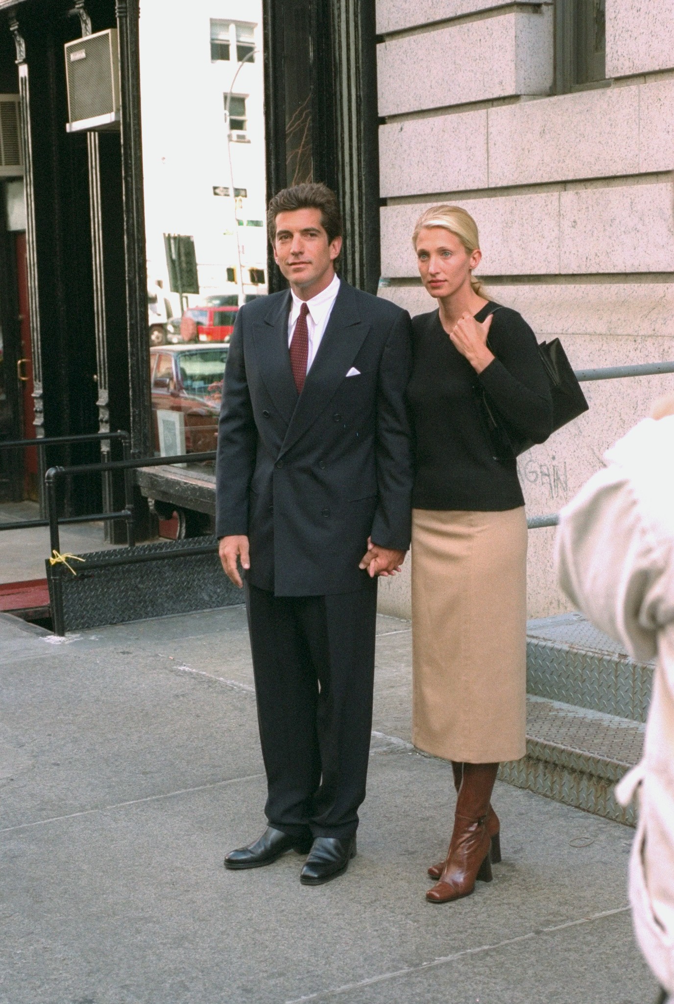 Carolyn Bessette-Kennedy wearing a black top, a khaki skirt, brown boots and a black bag; John F. Kennedy Jr. wearing a black suit and a red tie