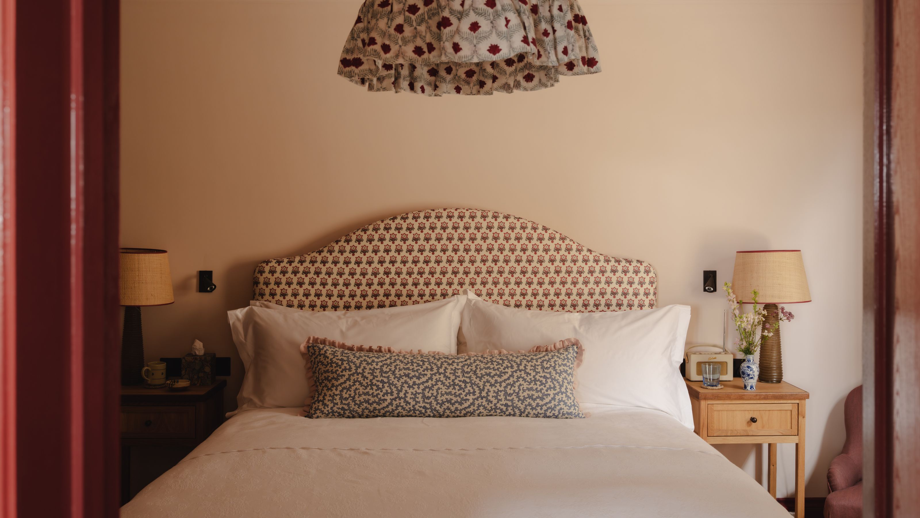 a pale pink bedroom with red door trim, a floral headboard, white bed linens and a patterned frilly lampshade