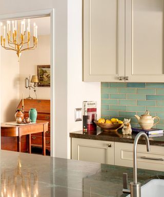A kitchen with ream cabinets, a turquoise blue tiled splashback, a black granite surface with a fruit bowl and books, and a larger black granite surface with a sink and a silver fridge to the left