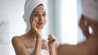 woman applying skincare in her bathroom with a towel on her head