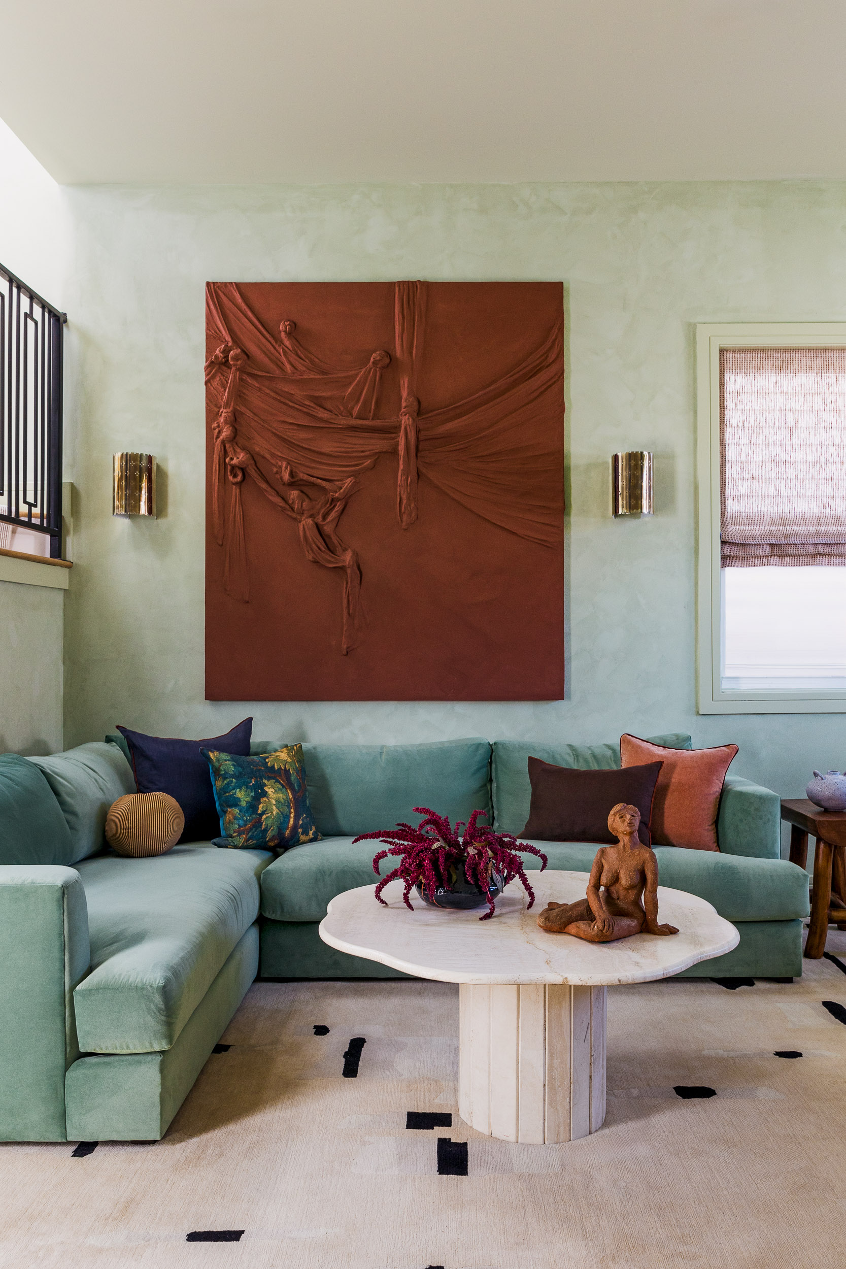 living room with mint green walls, mint-blue L-shaped sofa, orange draping artwork, round marble table with statue on it, and beige rug with black dots