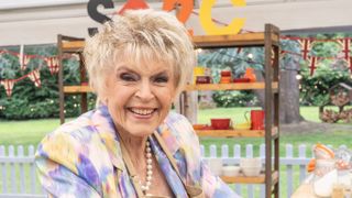 Gloria Hunniford wearing a pastel patterned top and smiling for the camera as she bakes in the Bake Off tent