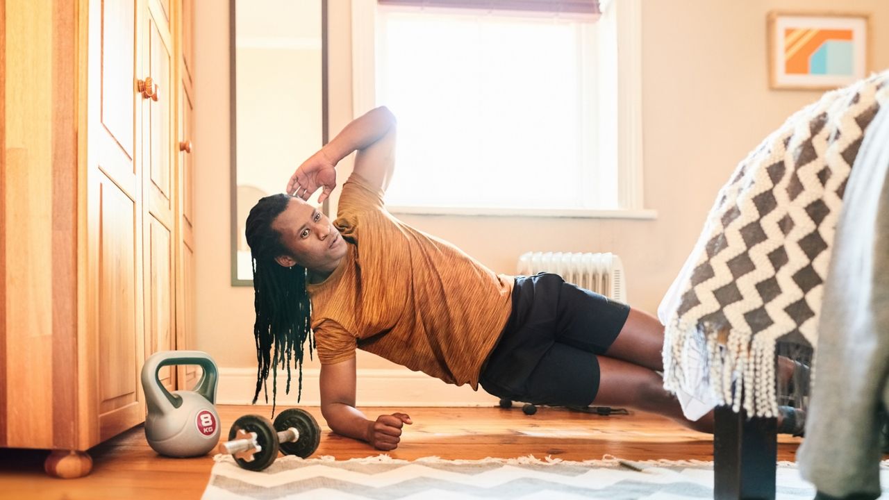 A man performs a side plank in a bedroom. He faces the camera, with his body held straight, weight balanced on his elbow and the side of one foot. Next to him is a wardrobe a kettlebell and a dumbbell. In front of him we see the corner of a bed.