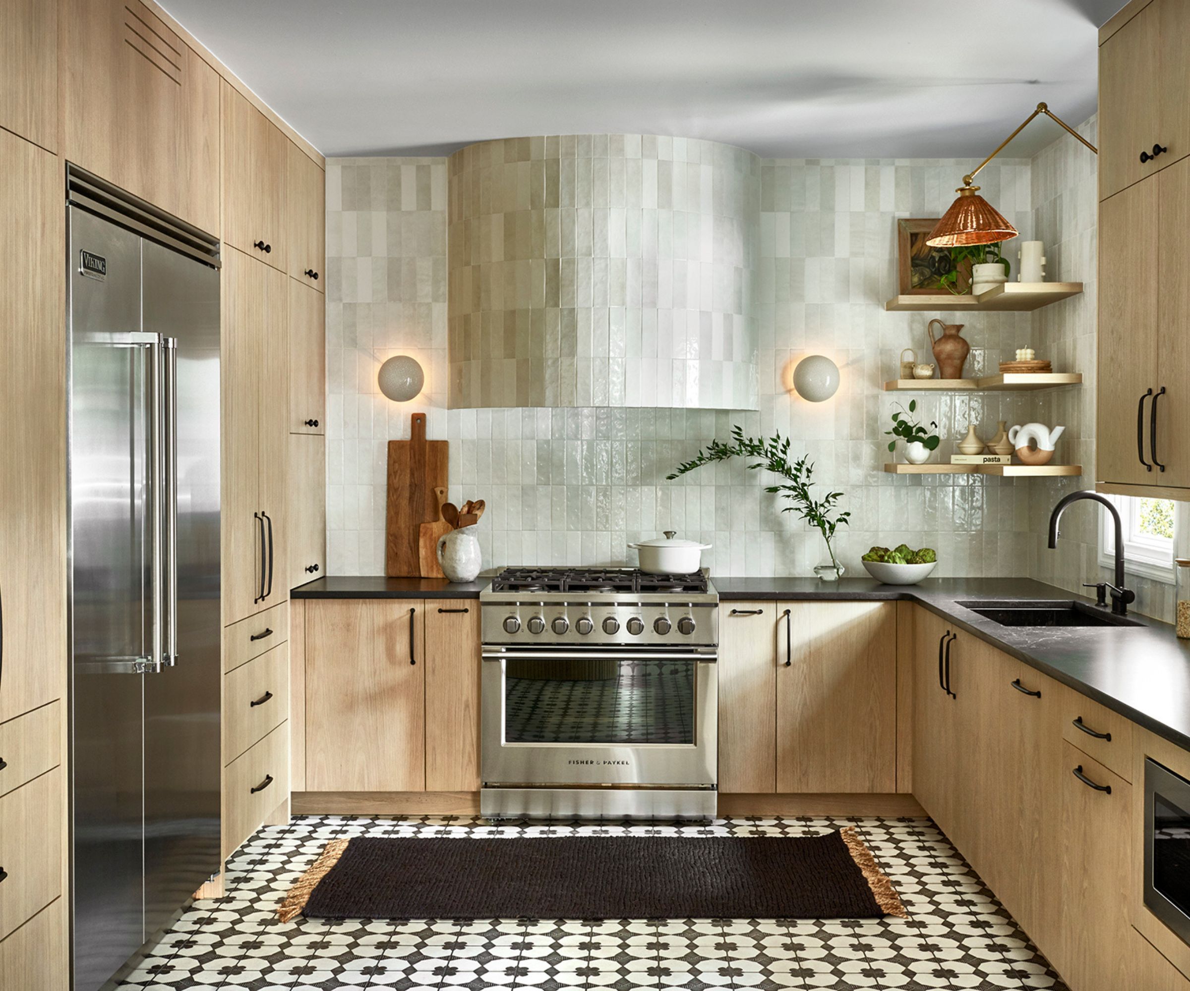 A wooden U-shaped kitchen with stainless steel appliances and an off-white zellige tile backsplash and matching curved range hood