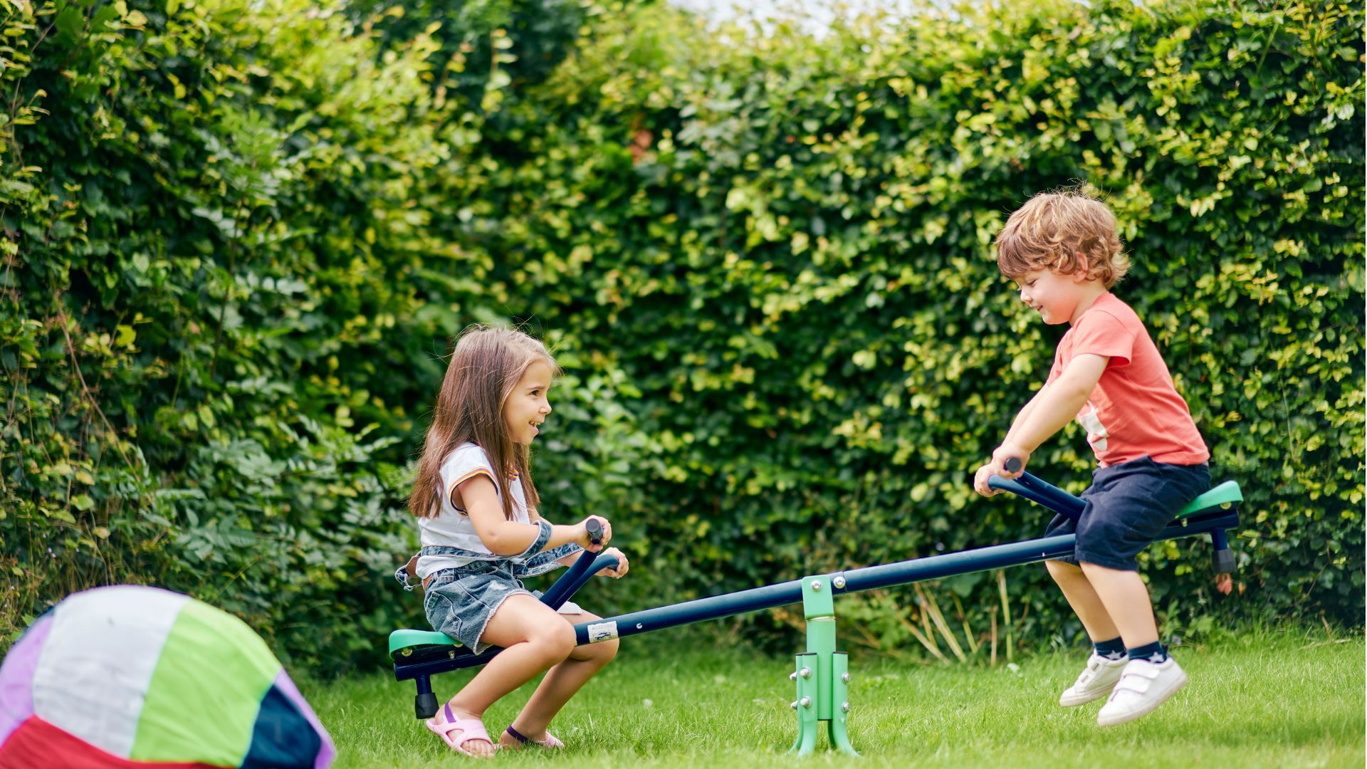 A young girl and boy play on a seesaw in a garden.