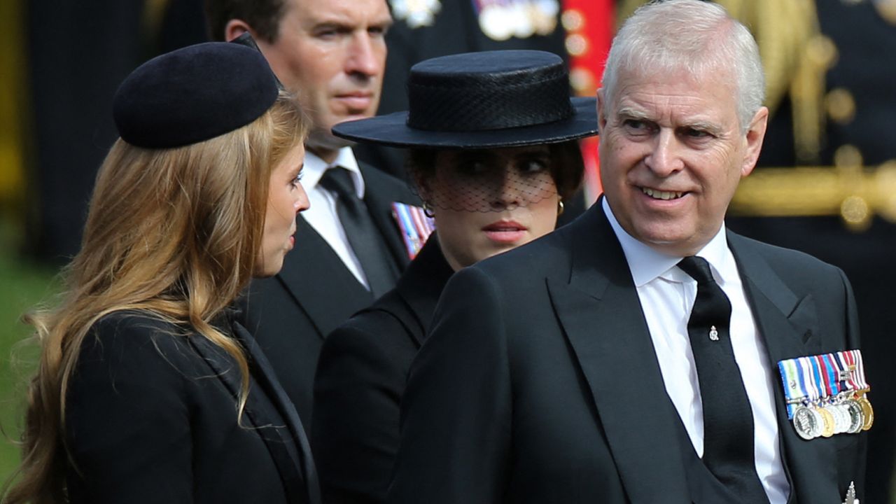 Princess Beatrice and Princess Eugenie wearing black hats and coats standing next to Prince Andrew who is smiling and wearing a black suit with military medals