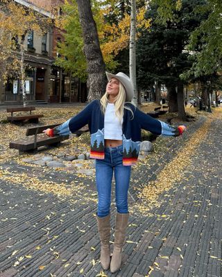 Bridgette wearing skinny jeans, knee-high boots, a graphic cardigan, a white cropped tee, and a cowboy hat.