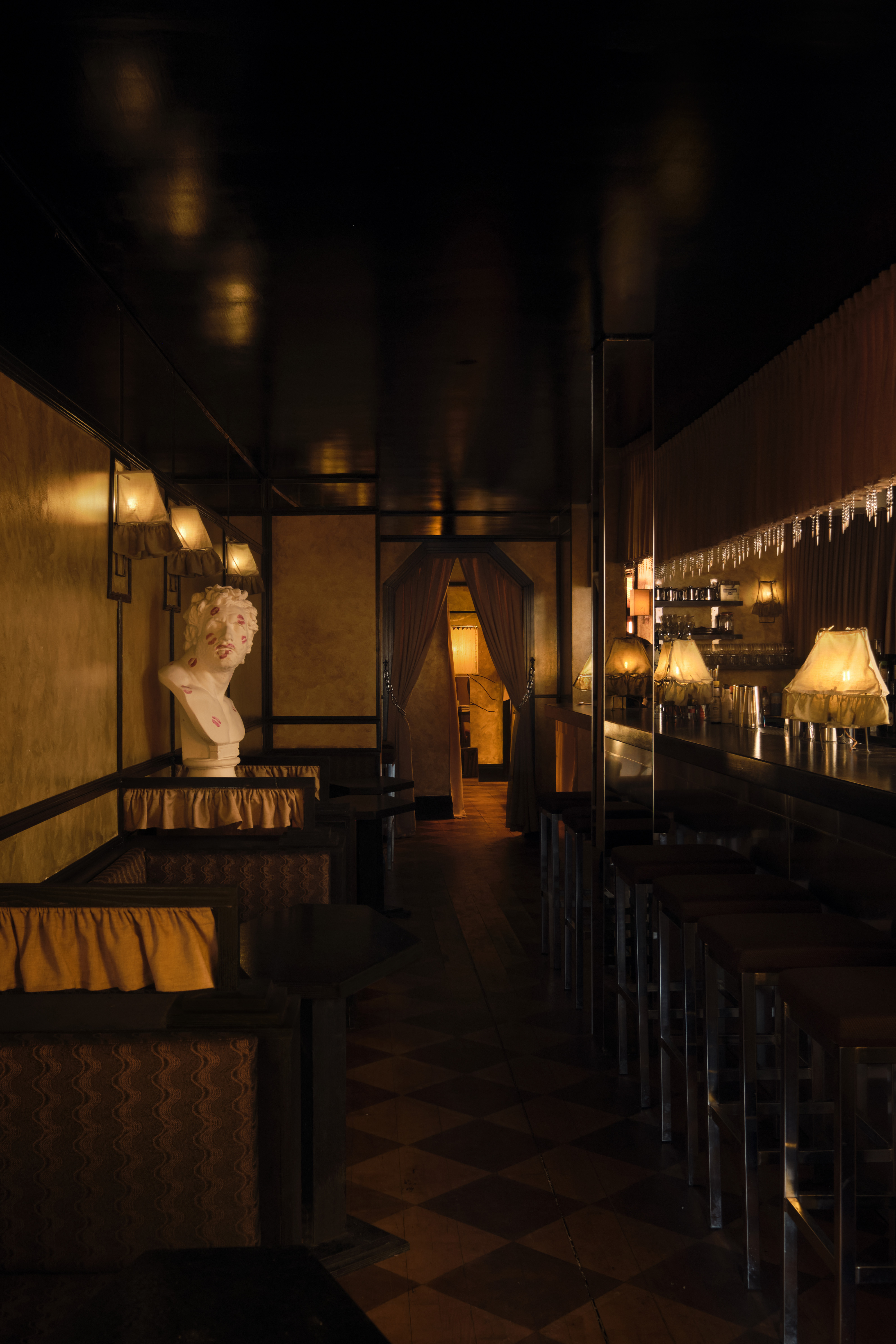 An amber-lit nightclub features retro-inspired furniture echoing the atmosphere of the 20th century, marble busts, plush fabrics, and wall murals.