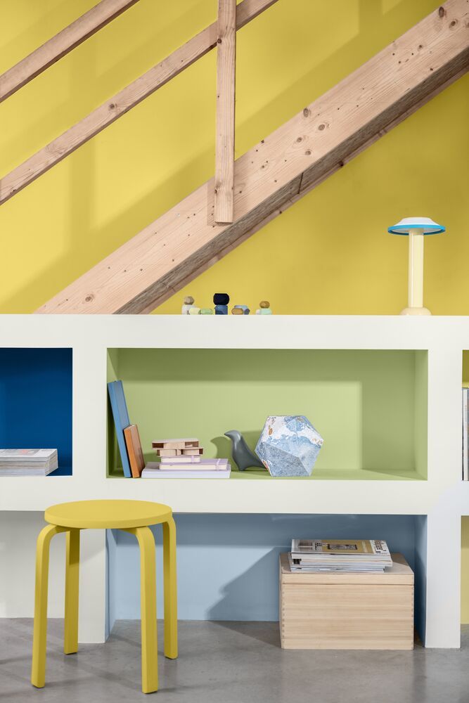 Image of a wooden staircase against a yellow wall. There is a low, white shelving unit with the inside painted in green, light blue, and dark blue. A small yellow stool is next to the shelving unit.