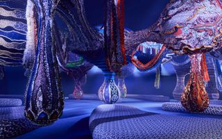 A surreal, large-scale textile installation features intricately woven, mismatched patches of fabric as well as beads, combined into a colossal and creature-like sculpture.