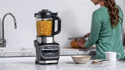 A Ninja Foodi Cold & Hot Blender making soup while a woman slices bread in the background