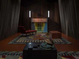 Inside the Luca Guadagnino Salone del Mobile 2022 installation featuring a dark room with a dark table and object in the foreground, a red reclining chair in the distance, a multi-shaped rug and a closed fireplace.