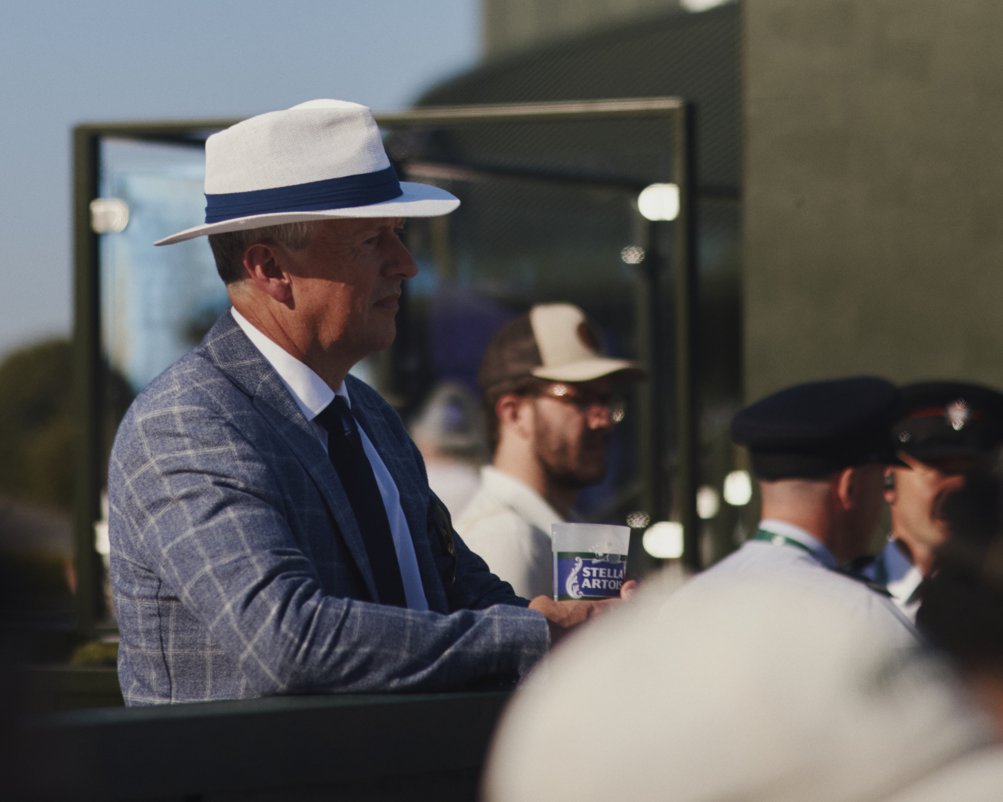 Man in hat, tie and suit jacket, holding a glass, watching Wimbledon tennis