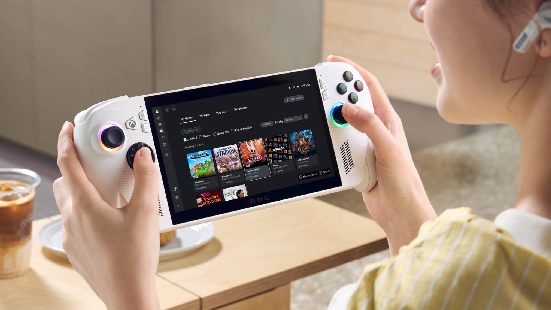 Player holding white Asus ROG Xbox Ally next to table with iced coffee on top.