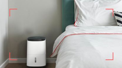Meaco dehumidifier next to bed with white linen in a light grey bedroom to support an expert guide on What you need to know before buying a dehumidifier