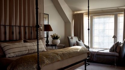 A cozy neutral bedroom with warm white walls, a large window with a bench seat and beige curtains, carpeted floor, a four-poster canopy bed with layered neutral bedding.
