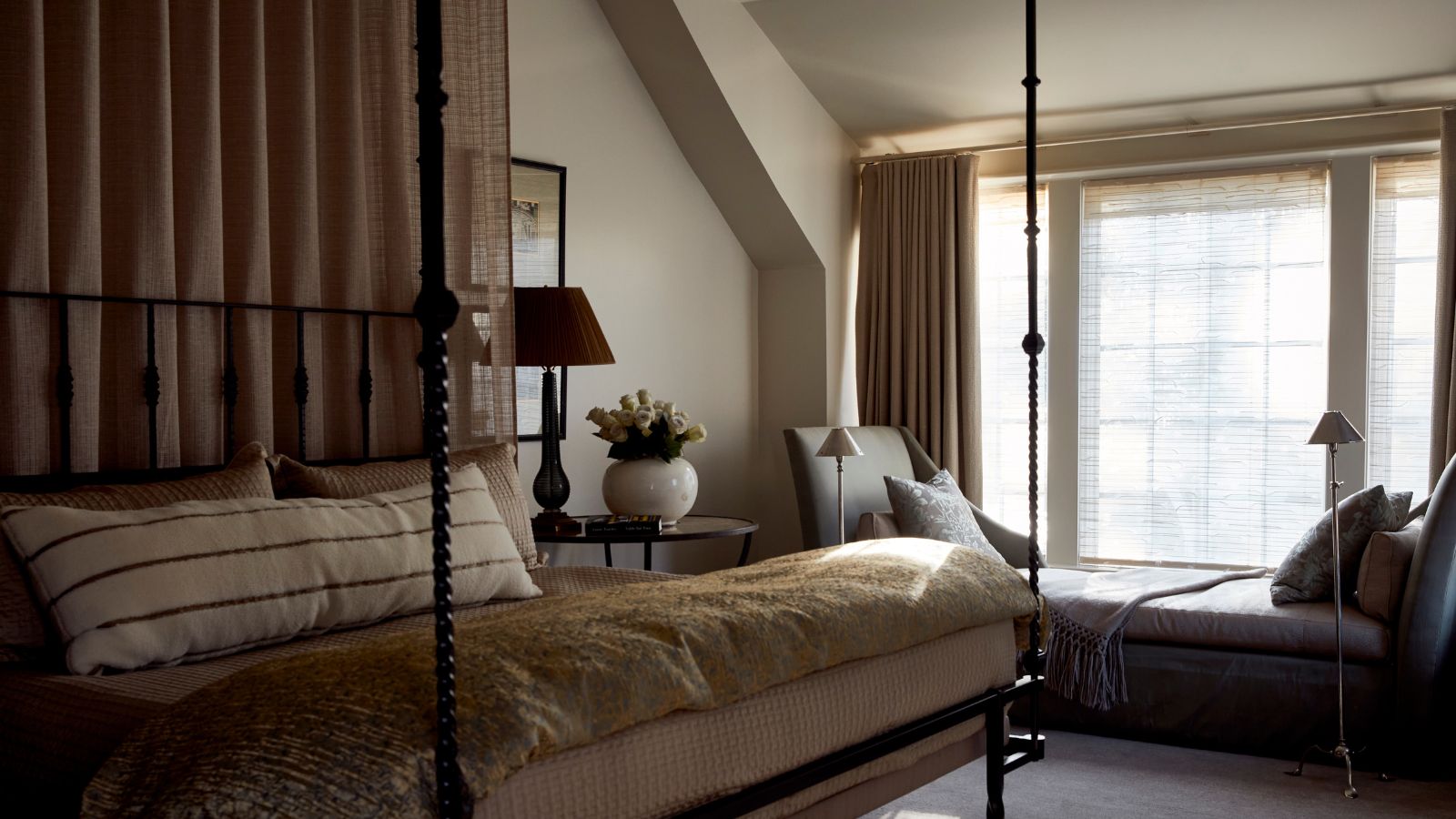 A cozy neutral bedroom with warm white walls, a large window with a bench seat and beige curtains, carpeted floor, a four-poster canopy bed with layered neutral bedding.