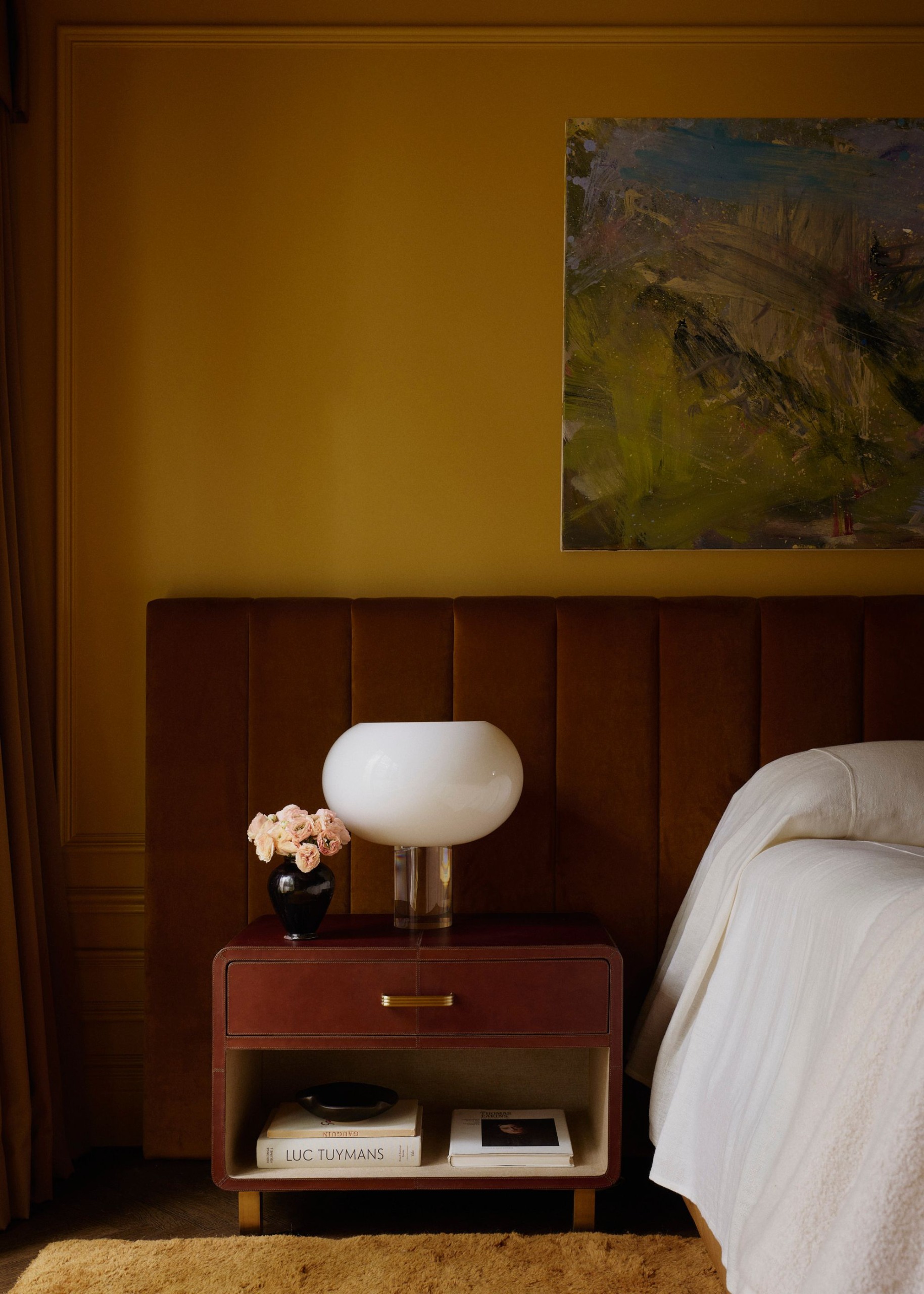 A moody bedroom with rich ochre walls, an extended headboard upholstered in brown velvet, a low wood midcentury nightstand, and white bedding. Blue and green abstract artwork on the wall.