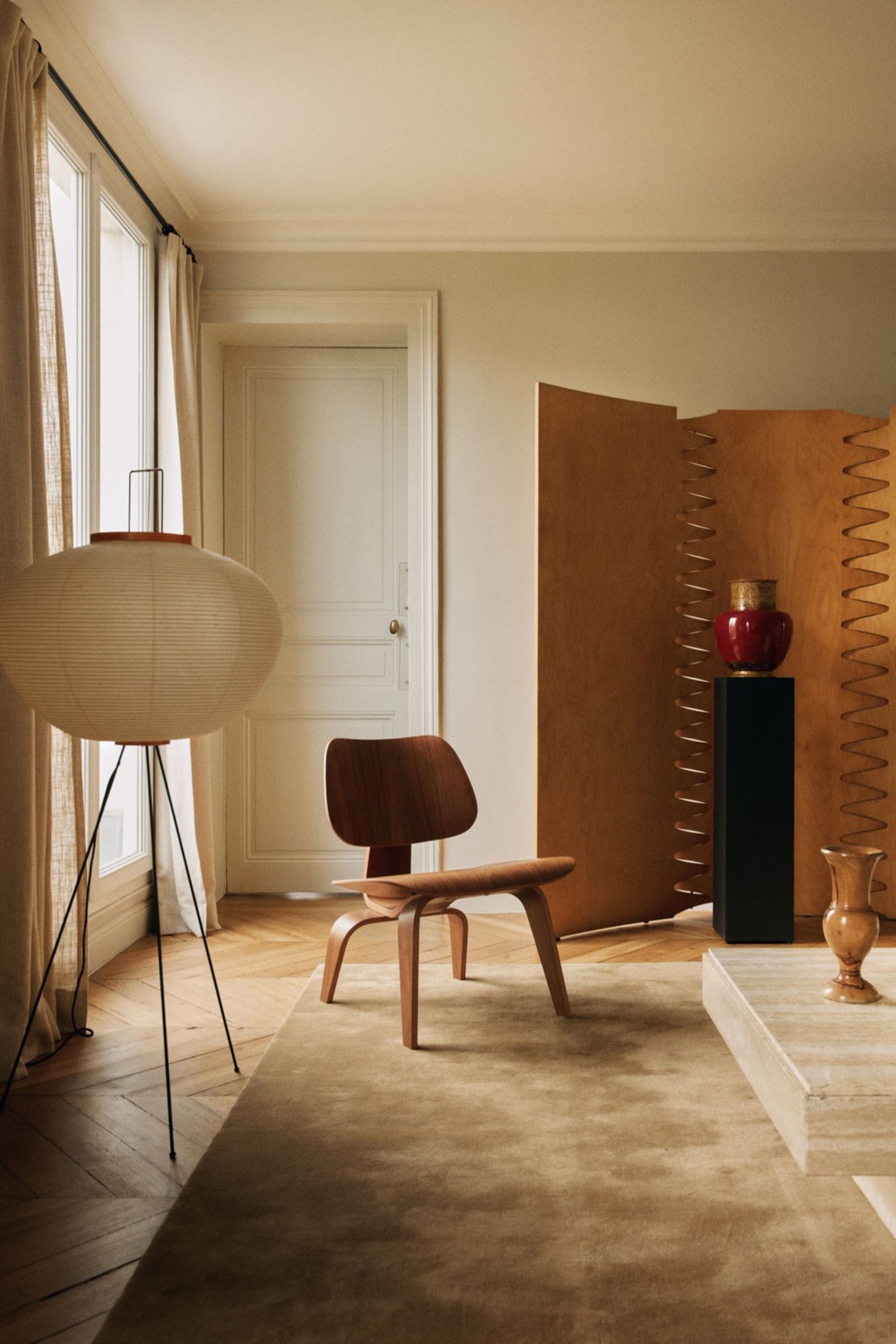 Living room with a wooden lounge chair, paper floor lamp, sculptural wooden screen, black pedestal, decorative vases and a beige rug on herringbone wood flooring
