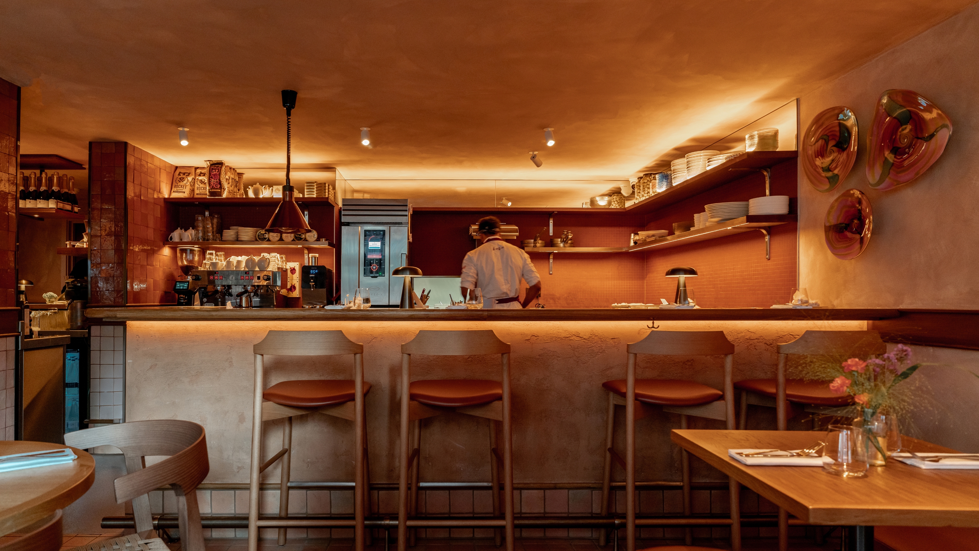 An orange-hued kitchen in a restaurant decorated with warm woods, a terracotta-hued series of furnishings, fresh flowers, and industrial-style lighting.