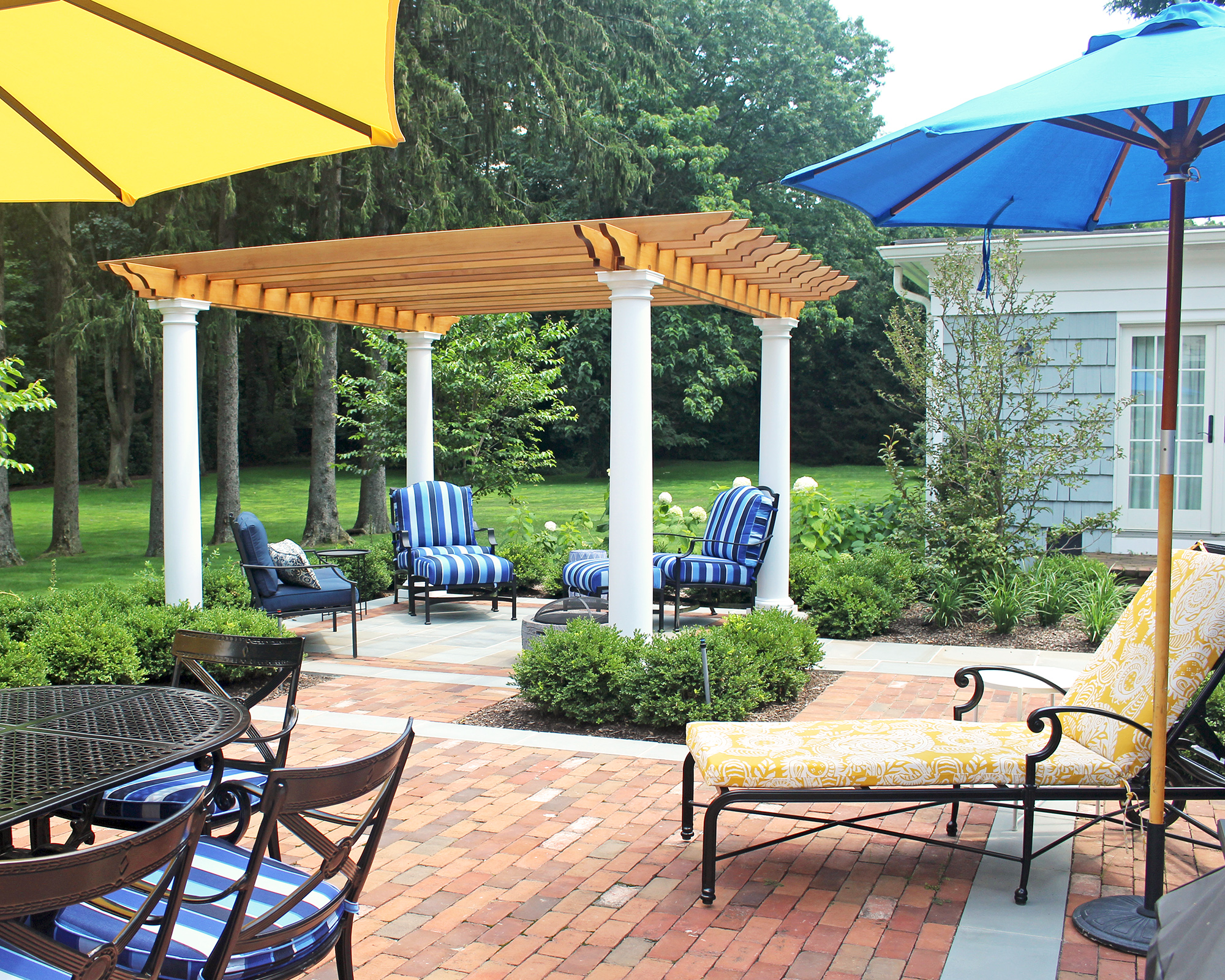Freestanding pergola with garden chairs on patio, with umbrellas, lounger and garden table with chairs, lawn and trees beyond