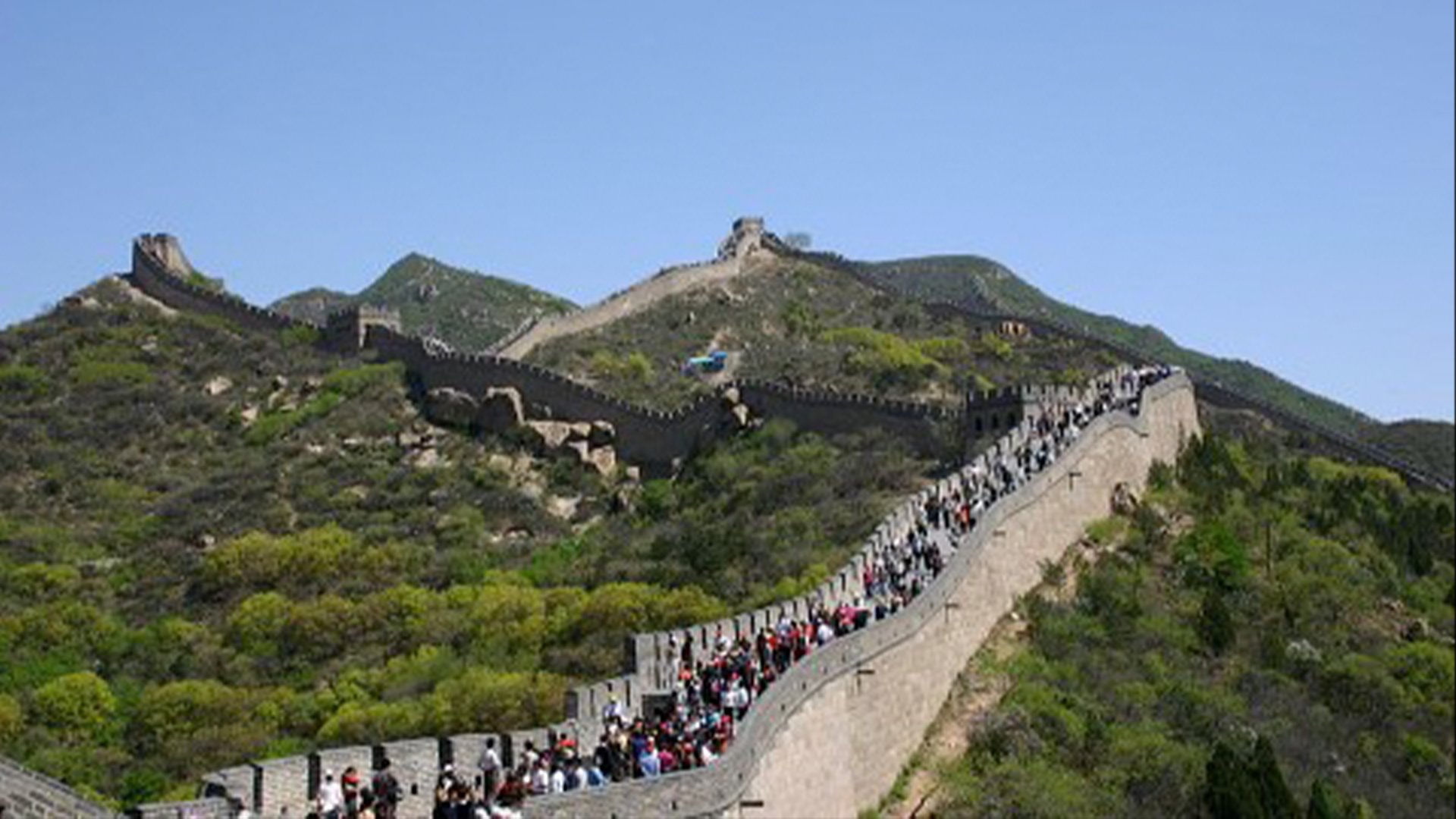 Un'ampia sezione della Grande Muraglia a Badaling si riempie di persone che camminano lungo la sua cresta.  La Grande Muraglia attraversa una catena montuosa verde sotto un cielo azzurro.