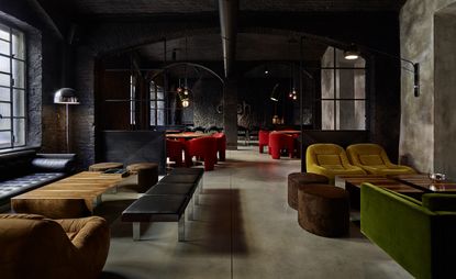 Interior view of Dash Kitchen featuring black and stone coloured walls and flooring, tables and black, brown, red, green and mustard coloured seating. Further back there is another area with tables, bright red chairs and pendant lights