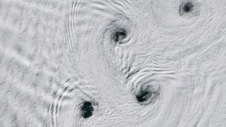 A white, snowy artic landscape has large swirling patterns with small holes or gaps at their center with what appears to be dark water underneath. 