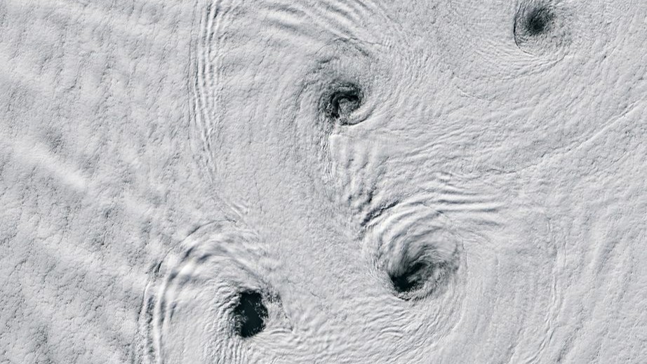 A white, snowy artic landscape has large swirling patterns with small holes or gaps at their center with what appears to be dark water underneath. 