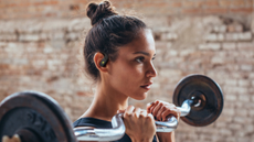A young woman with dark hair lifting weights in a gym while wearing JBL Endurance Zone headphones