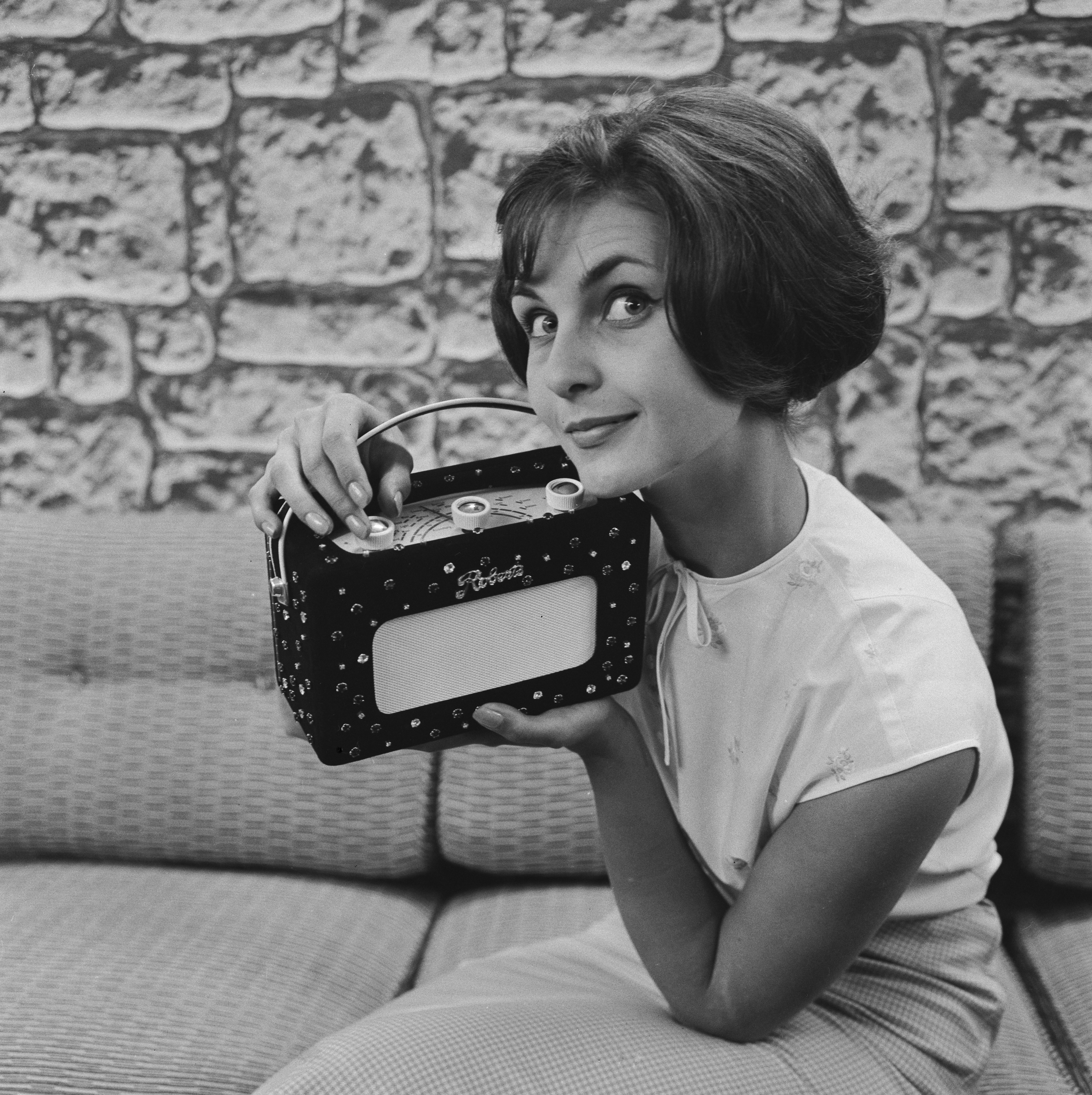 Black and white photograph of a woman with a bob, sitting on a sofa, holding a Roberts radio