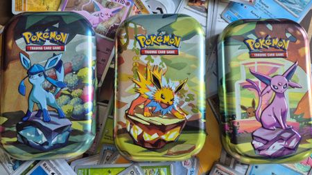Three mini Pokemon TCG Prismatic Evolutions tins on an array of cards
