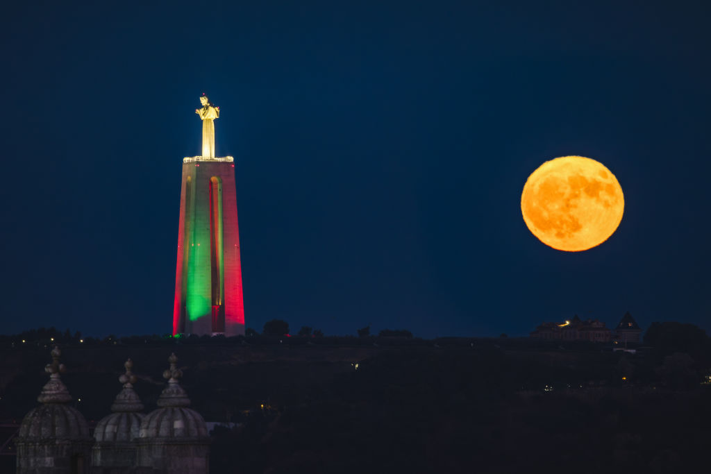 Duży posąg Chrystusa po lewej stronie jest oświetlony zielonym i czerwonym światłem, podczas gdy księżyc w pełni świeci jasno na niebie po prawej stronie posągu.