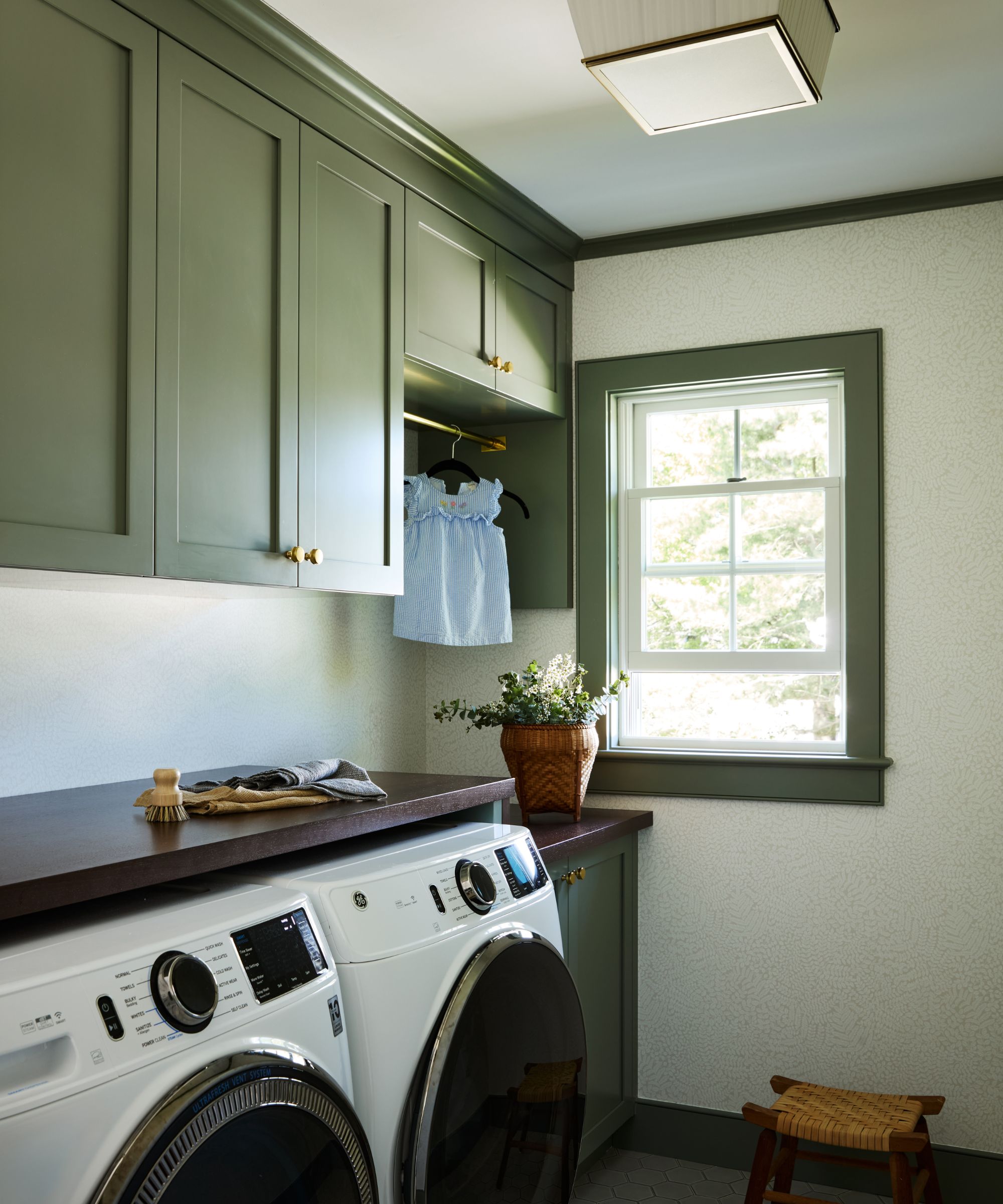 A laundry room with green cabinets, white wallpapered walls, green window trim, and brass hardware.