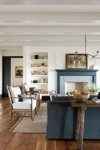 a traditional yet cozy living room design. The walls and ceilings are white and the floor is a maple-colored hardwood. There are two wooden accent chairs beside the slate-painted fireplace. Facing the fireplace is a slate blue fabric couch.