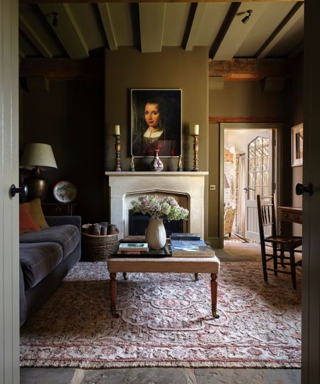 Country living room with beamed ceiling, dark brown painted walls, large fireplace with candlesticks and antique painting, Persian rug, velvet grey sofa and coffee table with books and a vase of flowers