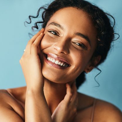 Elemis Dynamic Resurfacing Facial Wash - beautiful woman with her hand on her cheek smiling against a blue backdrop gettyimages:1181806328