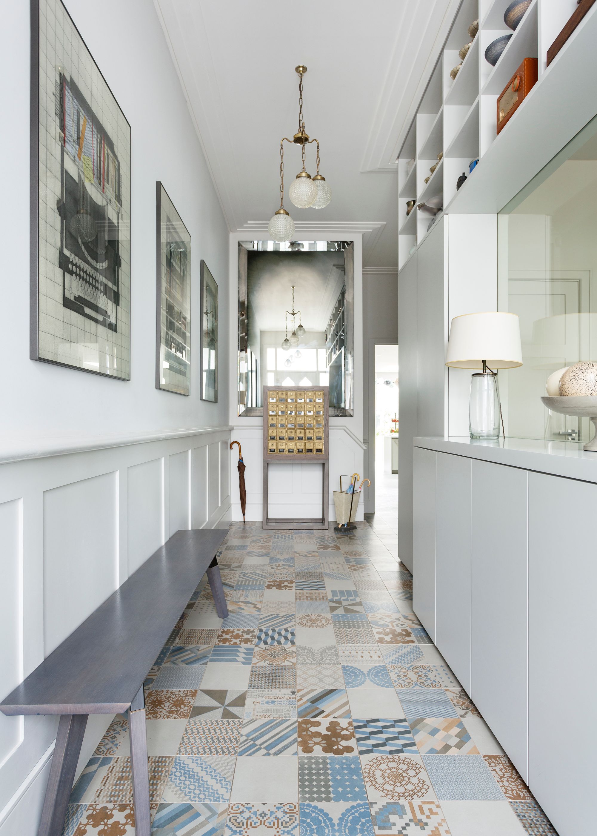 A modern hallway with earthy-colored patterned floor tiles, white walls, and light gray wainscoting, and a long gray bench.