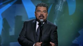 George Lopez performing stand-up comedy in a suit in front of a microphone.