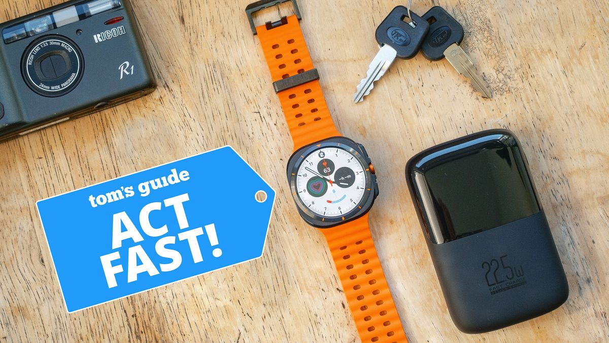 Samsung Galaxy Watch Ultra on an orange strap laying on a table next to some kets, a USB charger and a camera