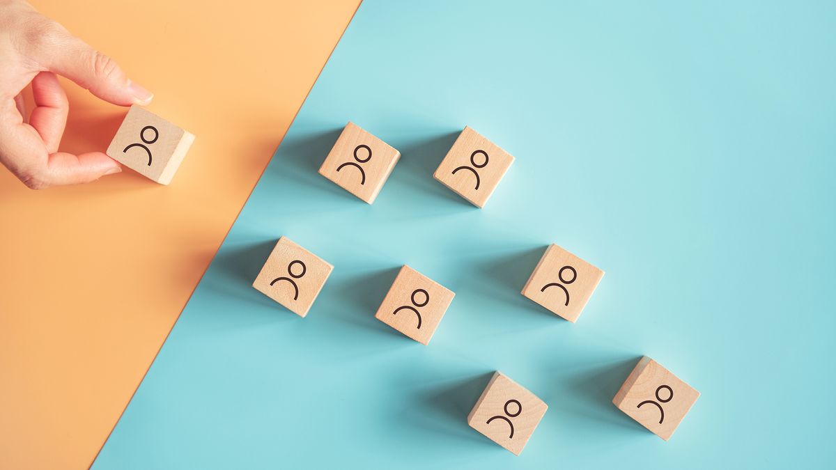 Eight wooden blocks with simple figures of people on them on a two-tone background, with a hand selecting one of them