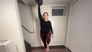 Jennifer Rizzuto performs a standing abs workout at home. She is marching on the spot. One hand holds a dumbbell overhead while the other swings at her side.