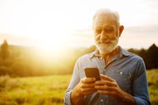A senior man stands outside looking at his phone.